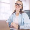 Junge Frau mit Brille sitzt lächelnd an einem Schreibtisch im Büro und arbeitet an einem Laptop.